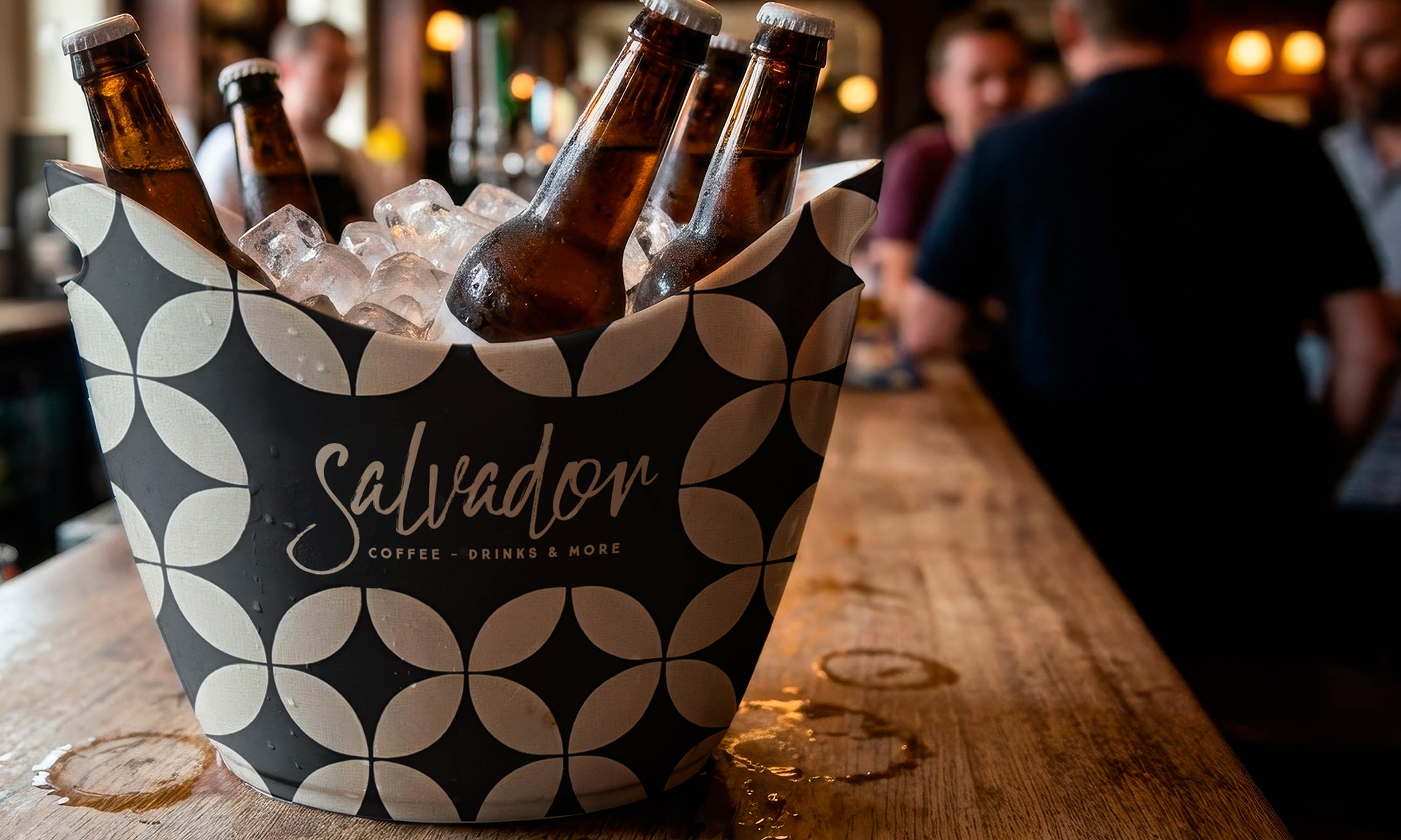 A wooden table features a Salvador Bar Athens bucket filled with six bottles of beer.