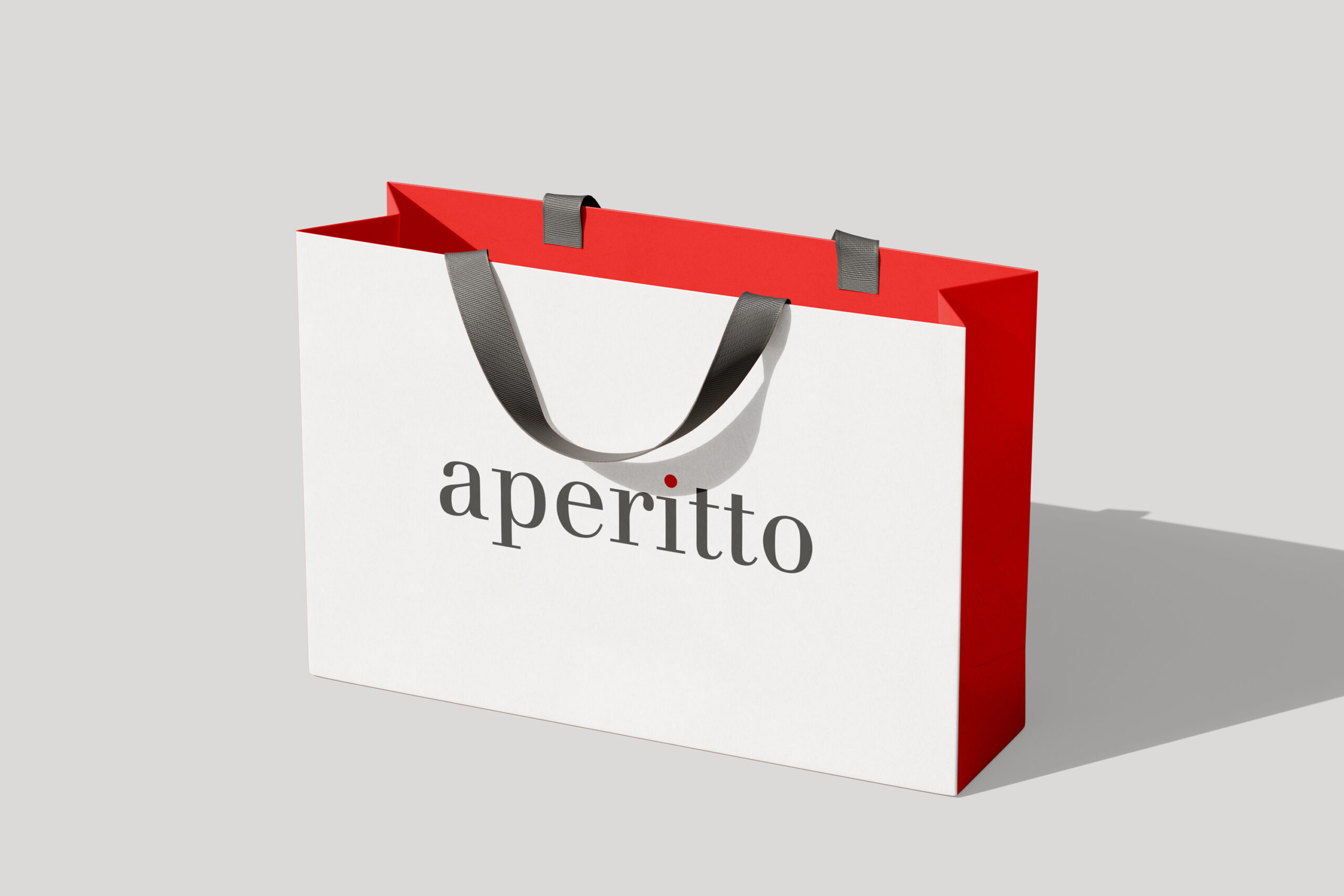 The image displays a white paper shopping bag with a vibrant red border at the top and red sides. Two dark gray, ribbed ribbon handles are attached to the inside of the red border. The word "aperitto" is printed in a dark gray, serif font centered on the front of the bag. A small, bright red dot is placed above the letter "i" in "aperitto." The bag is positioned at a slight angle, with its front facing the viewer and its right side visible. The background is a neutral, light gray, and a sharp shadow is cast by the bag to the right, indicating a light source from the upper left. The composition is clean and minimalist, focusing entirely on the shopping bag.