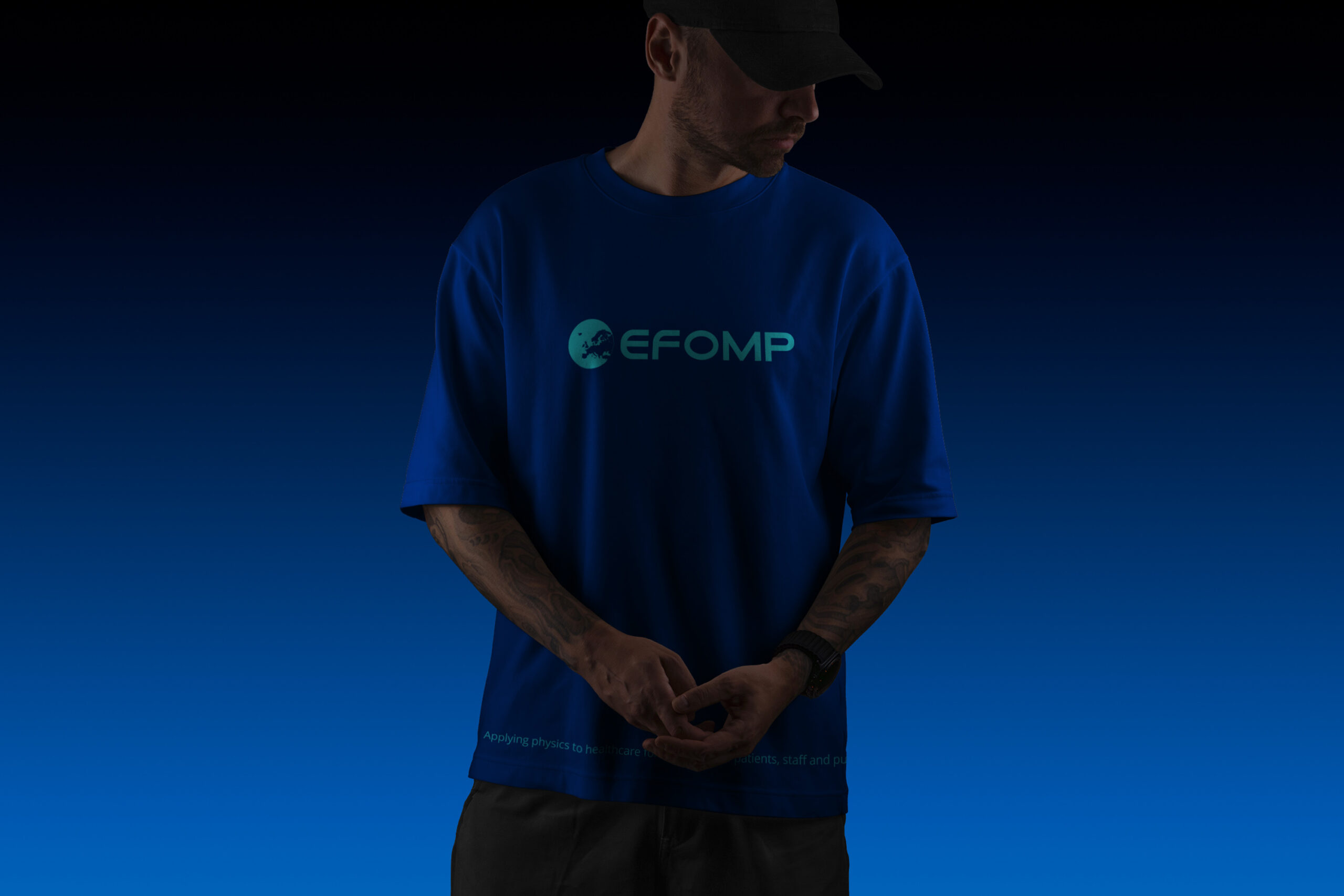 A man wearing a blue t-shirt with the EFOMP logo and text is pictured against a blue gradient background. The t-shirt also has text on the bottom hem.
