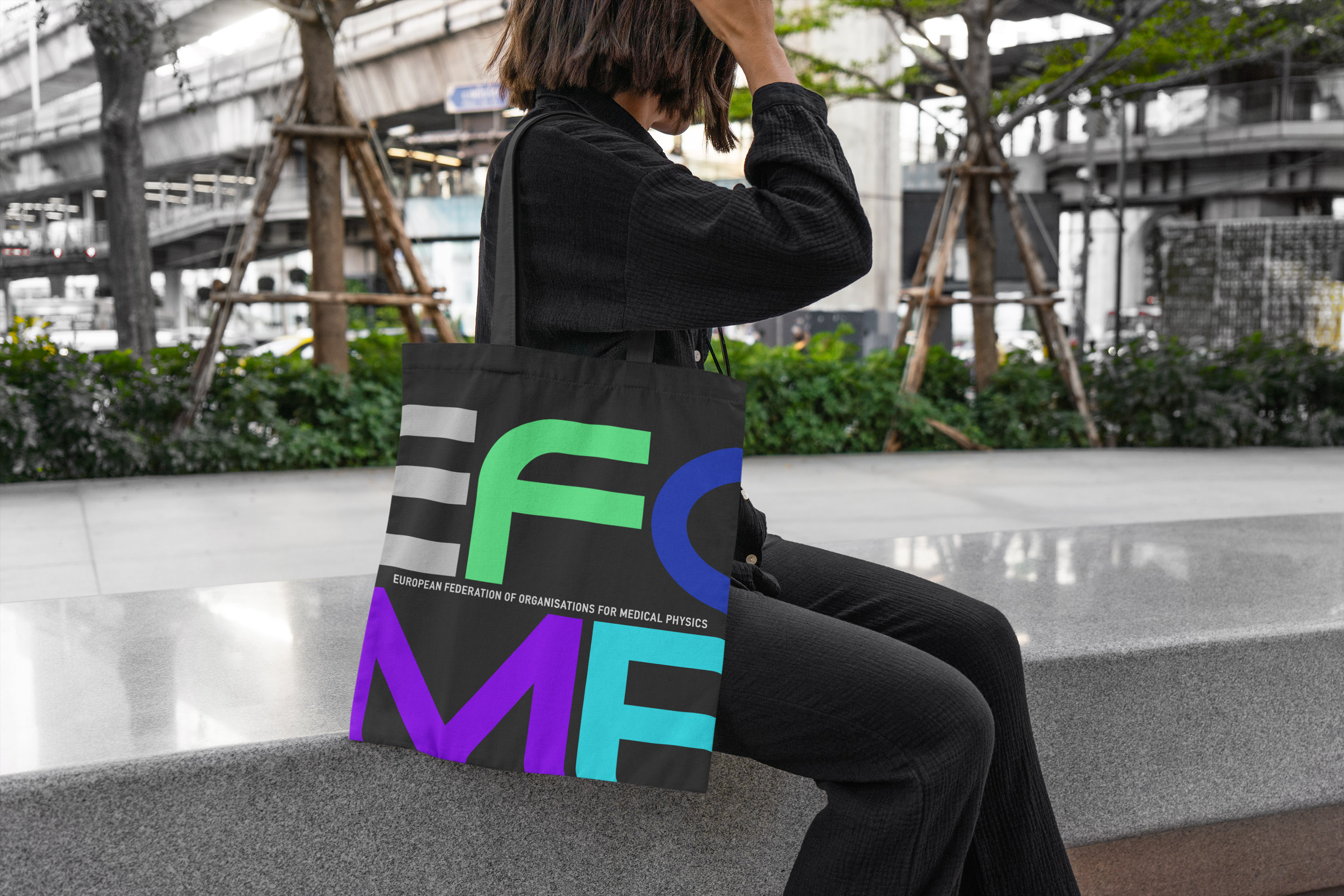 A person sits outdoors with a black tote bag featuring colorful lettering that spells "EFOMP". The bag also has text that reads "EUROPEAN FEDERATION OF ORGANISATIONS FOR MEDICAL PHYSICS".