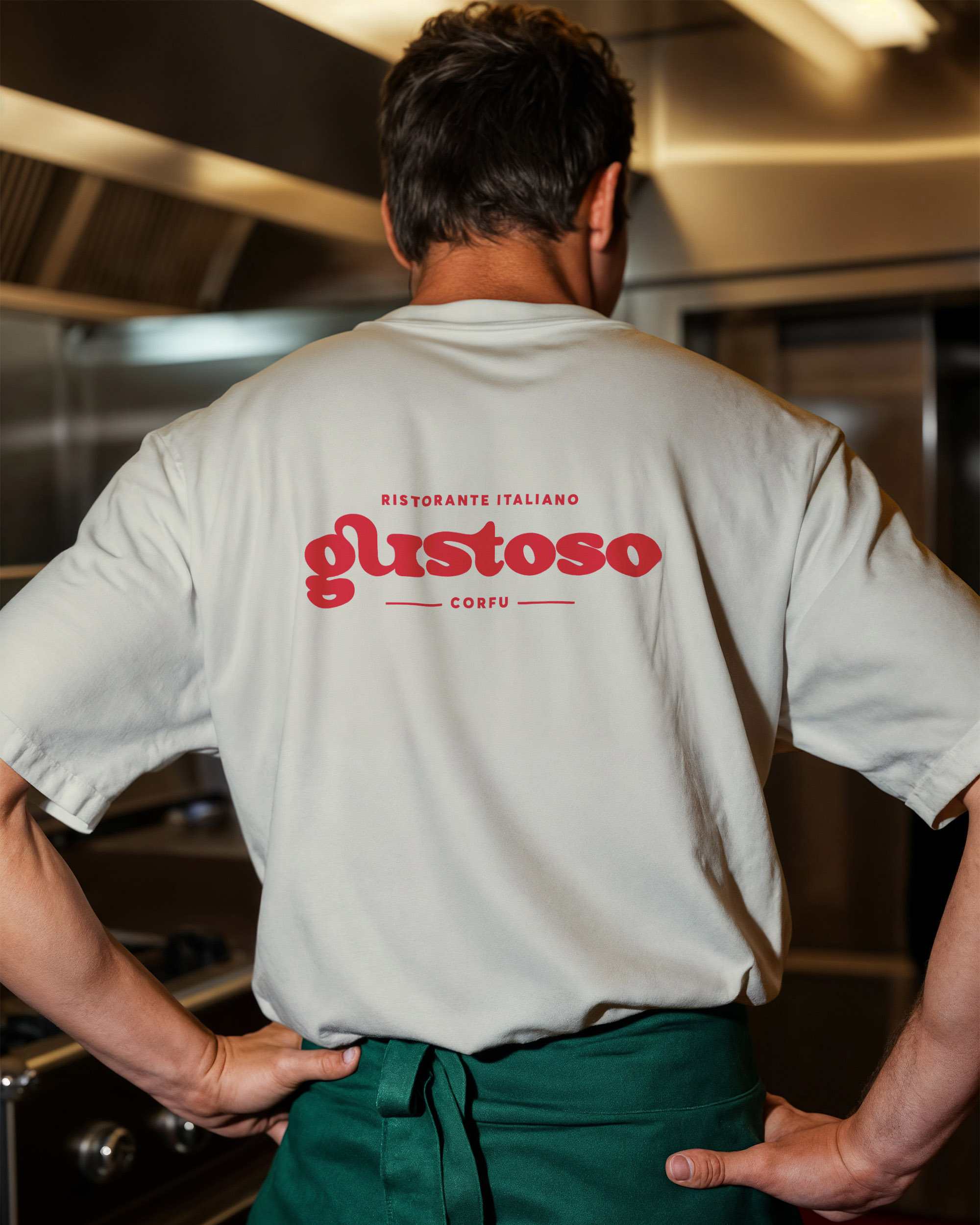 A man stands in a kitchen, wearing a white t-shirt with "Gustoso" printed on it, surrounded by cooking utensils.