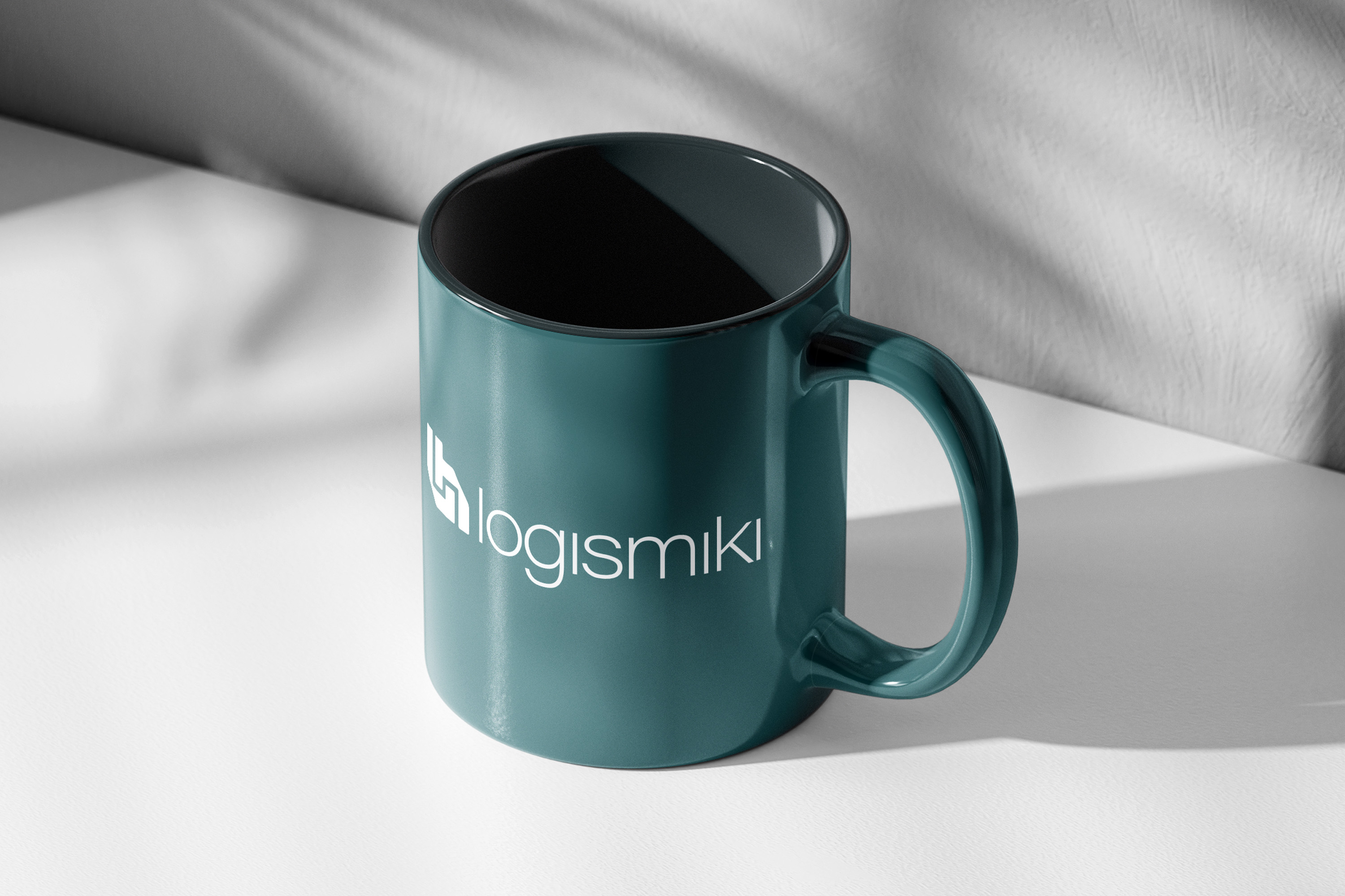 A teal mug with the white "logismiki" logo is placed on a white surface. Sunlight casts shadows of leaves on a textured gray wall behind the mug.