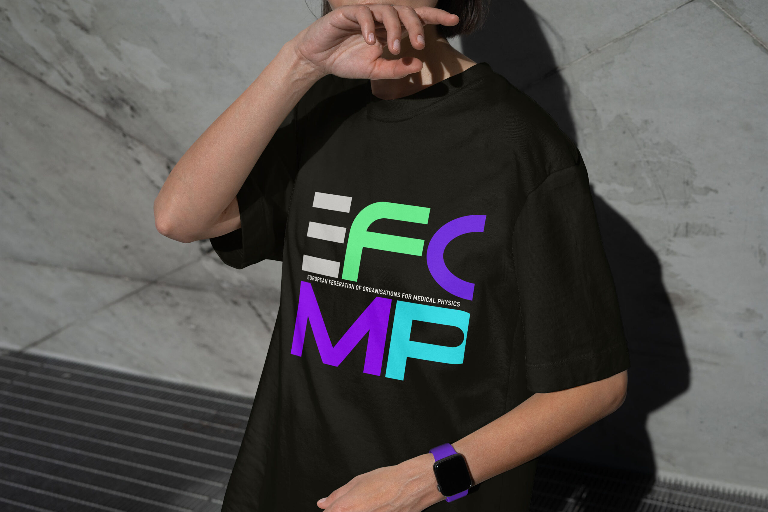 A person wears a black t-shirt with the colorful EFCMP logo and text. They are positioned in front of a grey concrete wall.